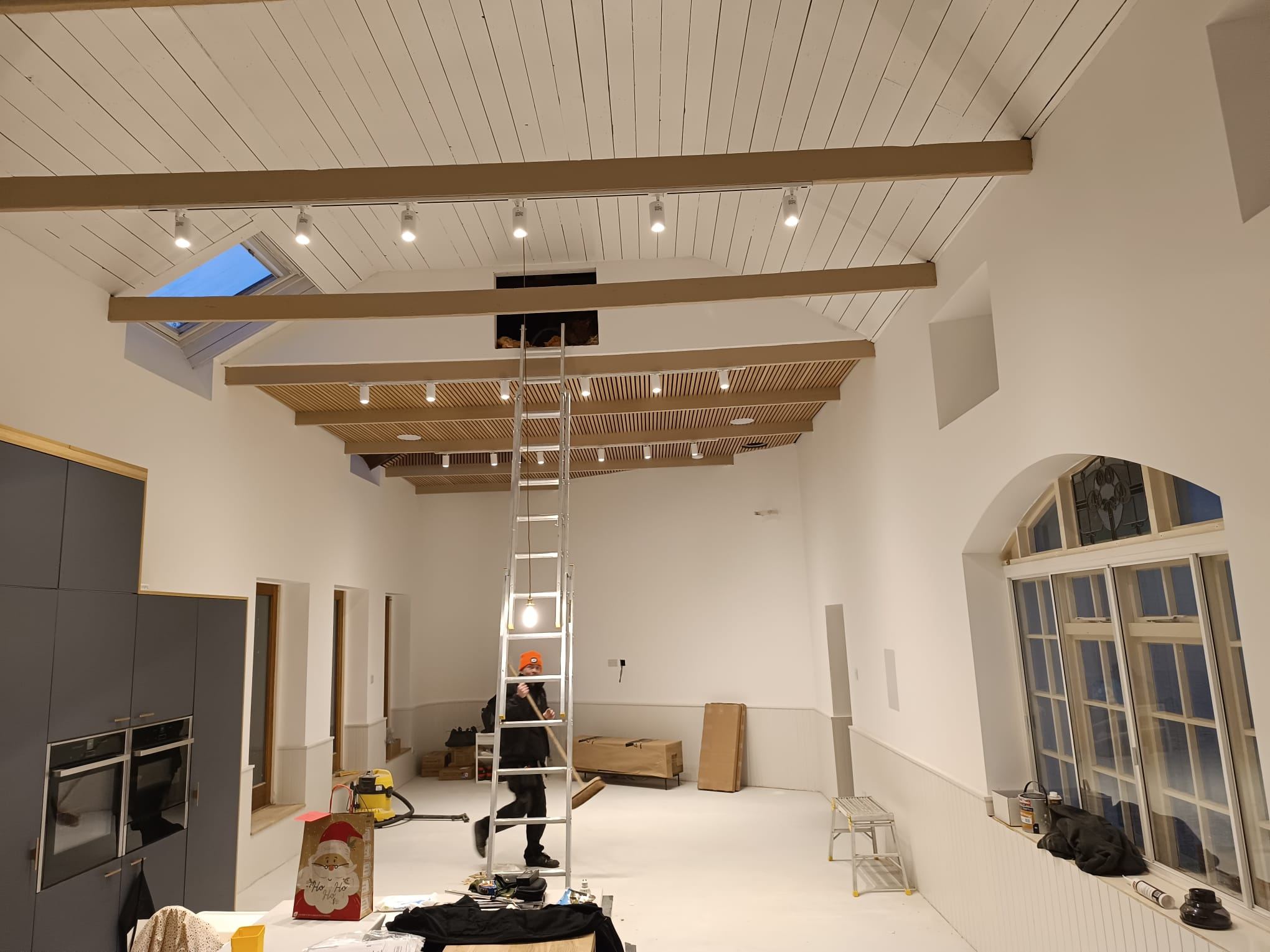 Hall conversion with track spotlights along timber beams and a slatted ceiling, an electrician on a ladder