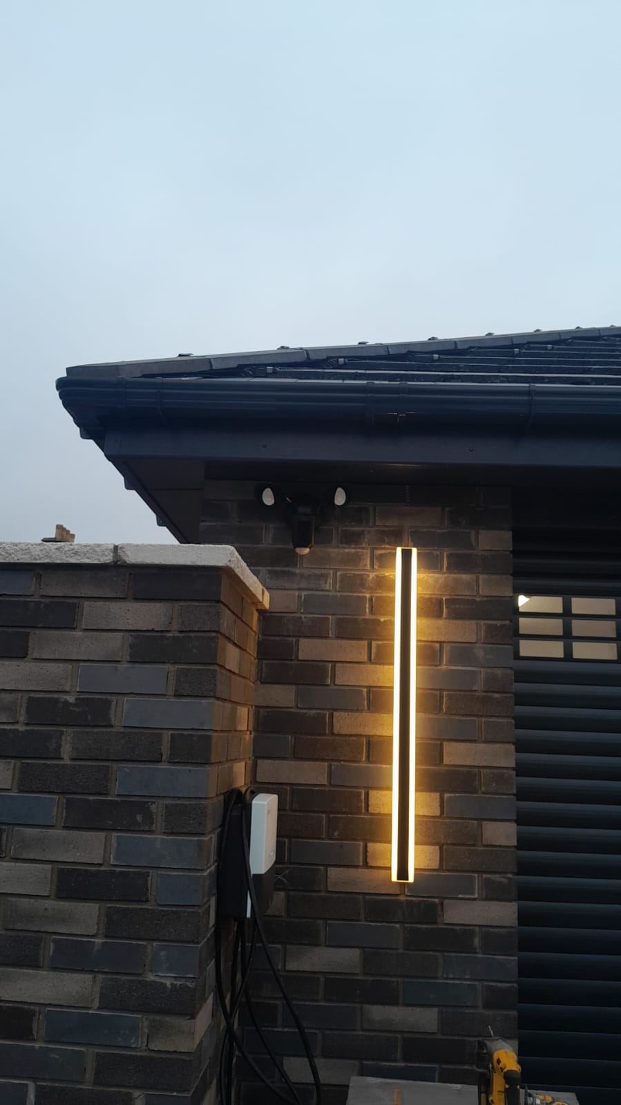 New dark-brick garage at dusk with an EV charger, a PIR floodlight and a warm linear wall light