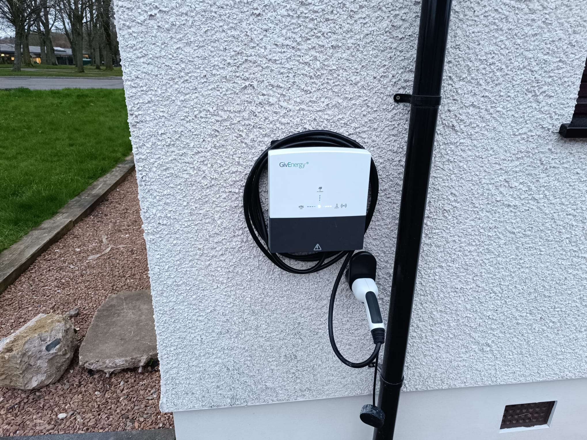 GivEnergy home EV charger with its tethered cable coiled, mounted on a white rendered wall beside a downpipe