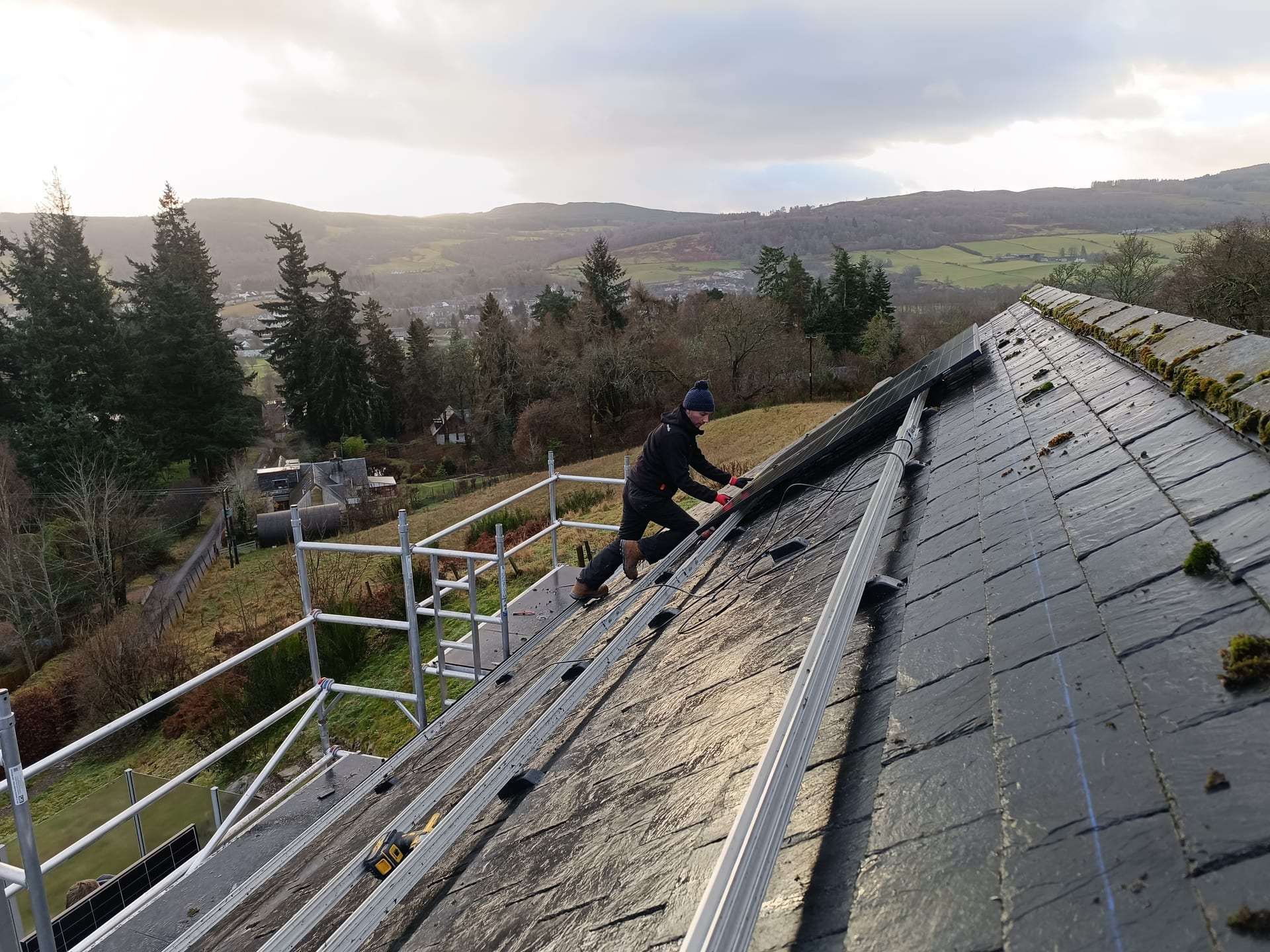 A Dual engineer fitting solar panels on a slate roof above Aberfeldy, hills in the distance