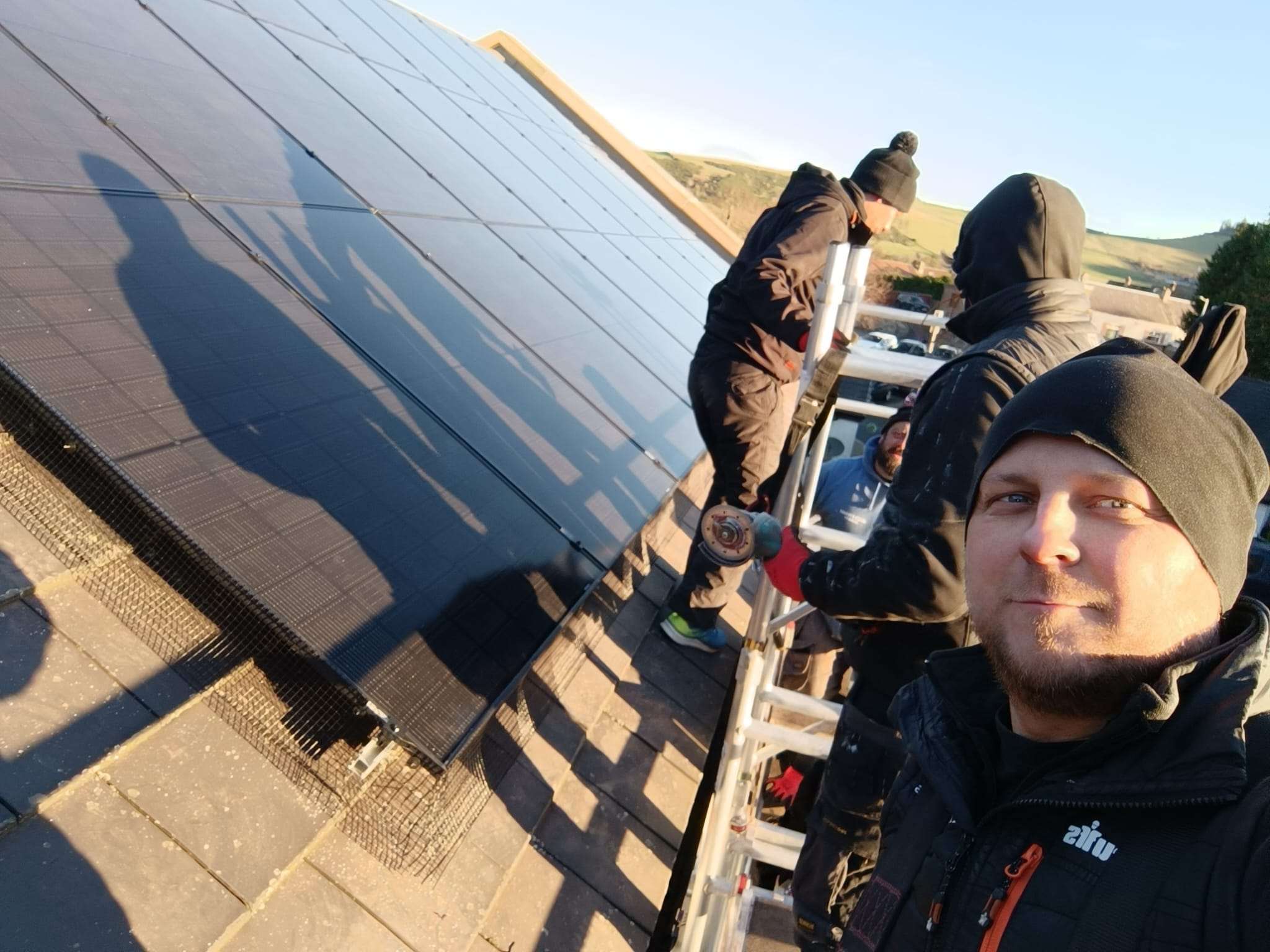 The Dual team on scaffolding beside a freshly fitted solar array, hills behind